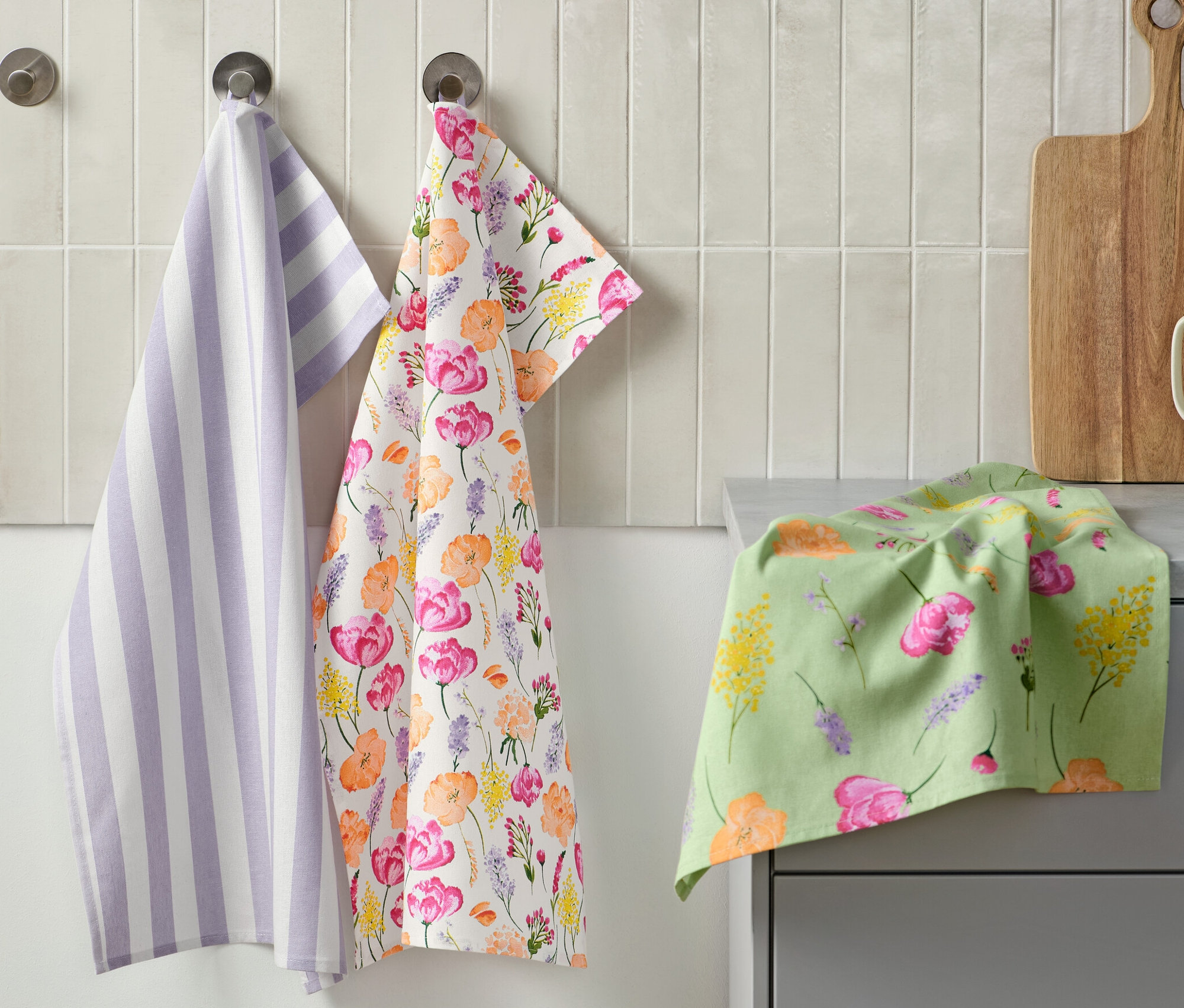 Na zdi visí tři utěrky: jedna fialovo-bílá pruhovaná, jedna s květinovým vzorem a jedna leží na pultu. Vedle je prkénko a hrnek s kuchyňským náčiním. Utěrky, 3 ks.