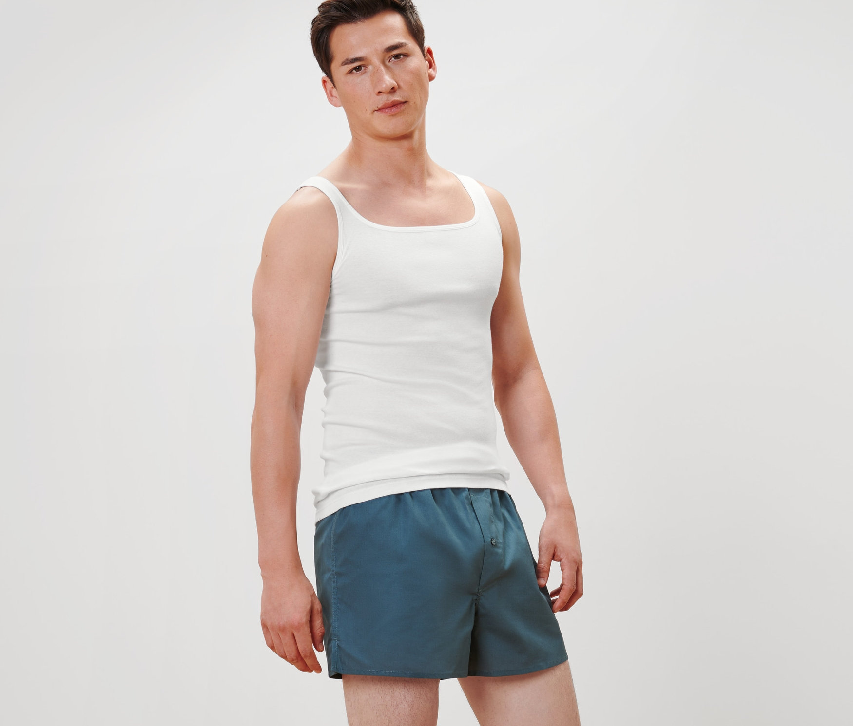 Muž stojí a má na sobě 2 bílá tílka z kvalitního, jemně žebrovaného úpletu a modré šortky.