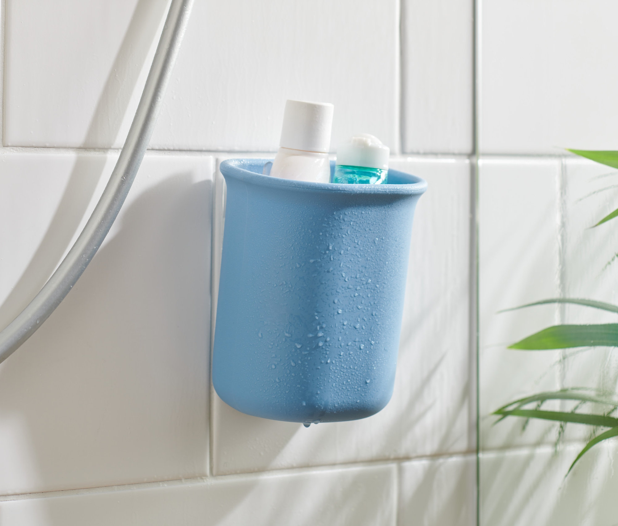 Modrý koupelnový organizér s bílým a modrým produktem uvnitř, připevněný na bílých dlaždicích vedle sprchové hadice.