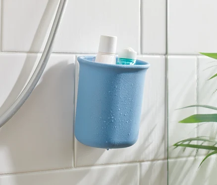 Modrý koupelnový organizér s bílým a modrým produktem uvnitř, připevněný na bílých dlaždicích vedle sprchové hadice.