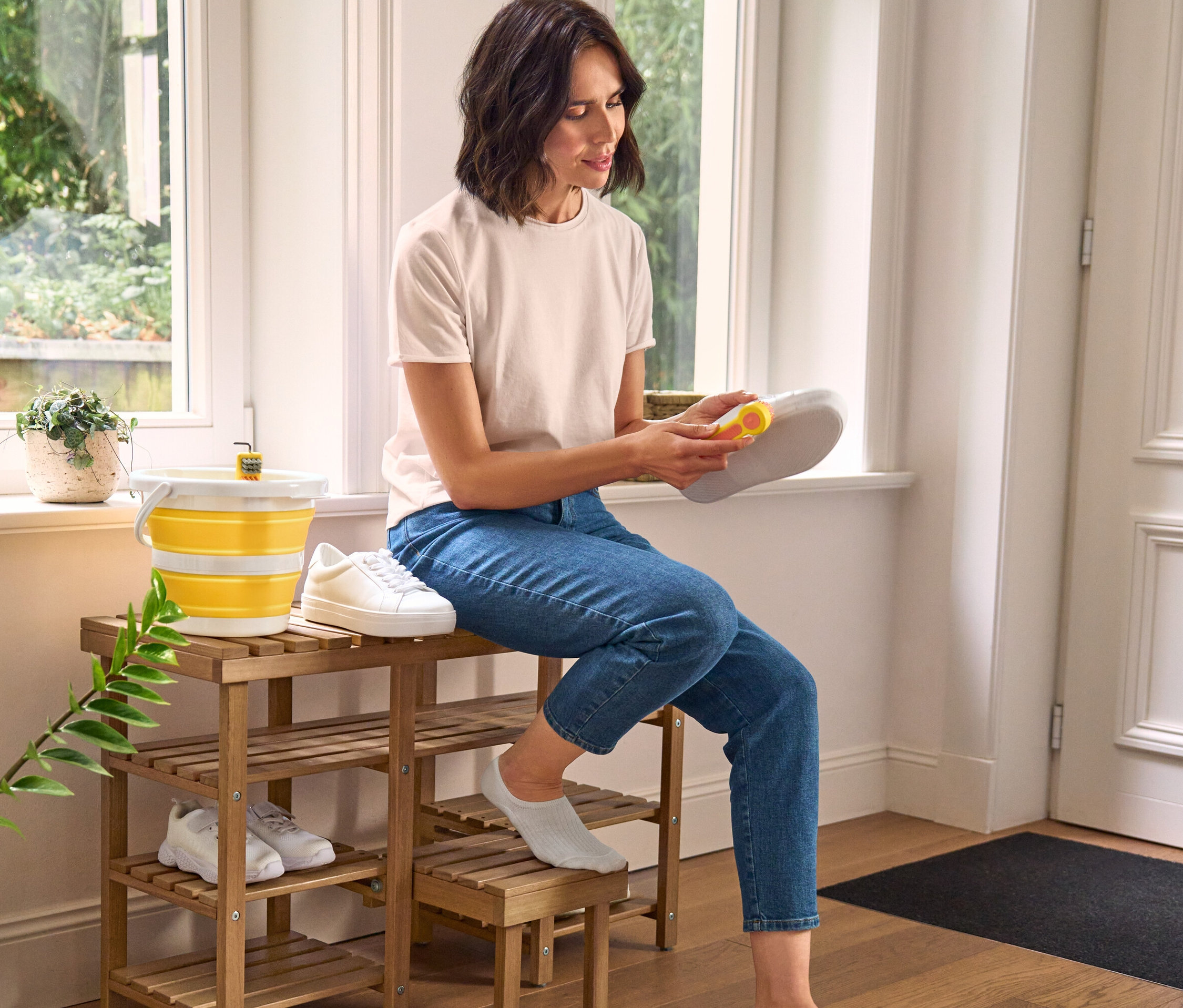 Žena čistí botu kartáčkem na boty 3 v 1, sedí na vysoké lavici s botníkem. Vedle ní stojí skládací úklidový kbelík a kartáč na boty.