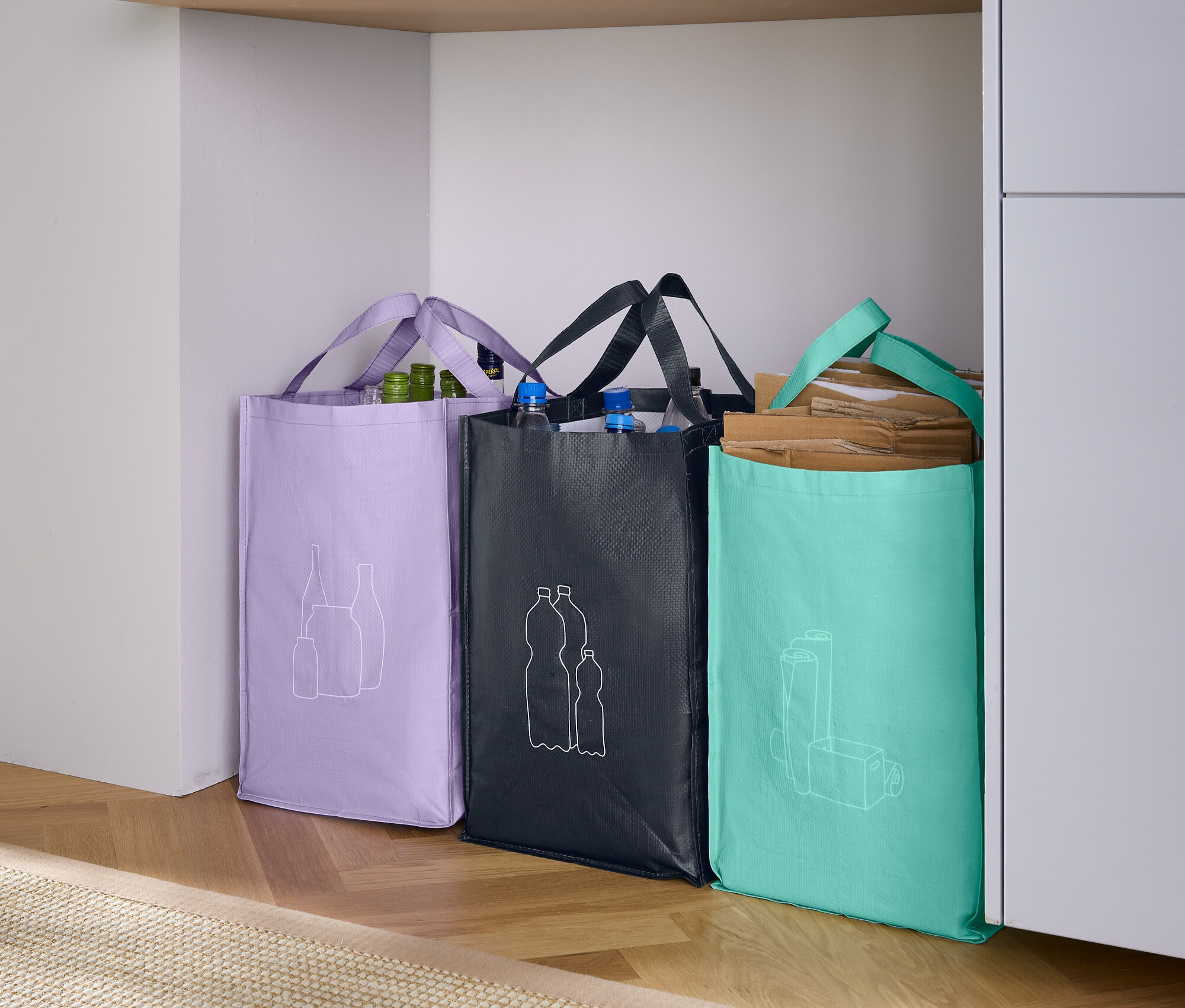 Tři nákupní tašky stojí vedle sebe: fialová s nákresem ovoce, černá s nákresem lahví a tyrkysová s nákresem sklenice.