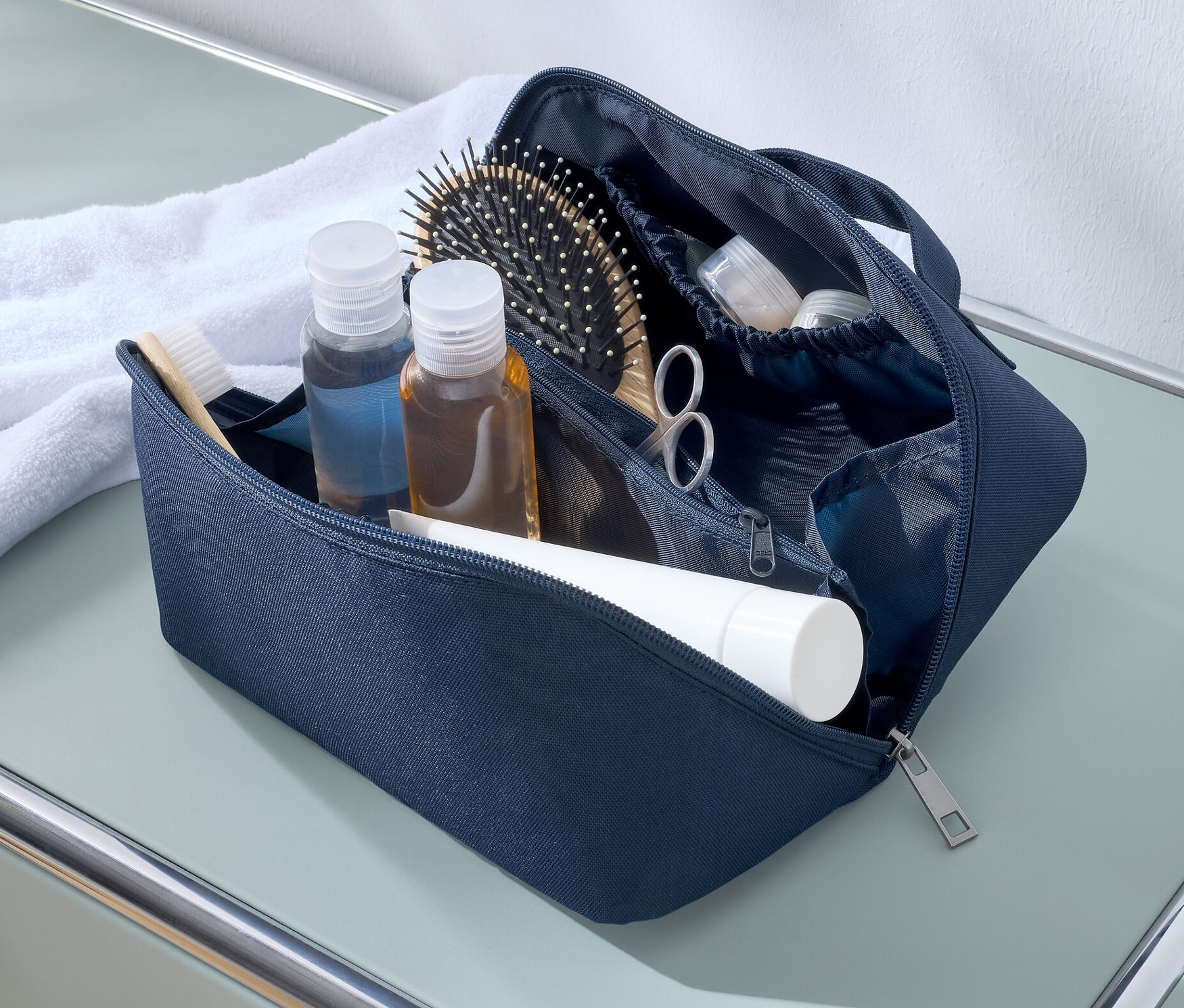 Modrá kosmetická taštička leží otevřená na stole vedle bílého ručníku. Uvnitř jsou cestovní lahvičky, kartáč na vlasy, nůžky a tuba.