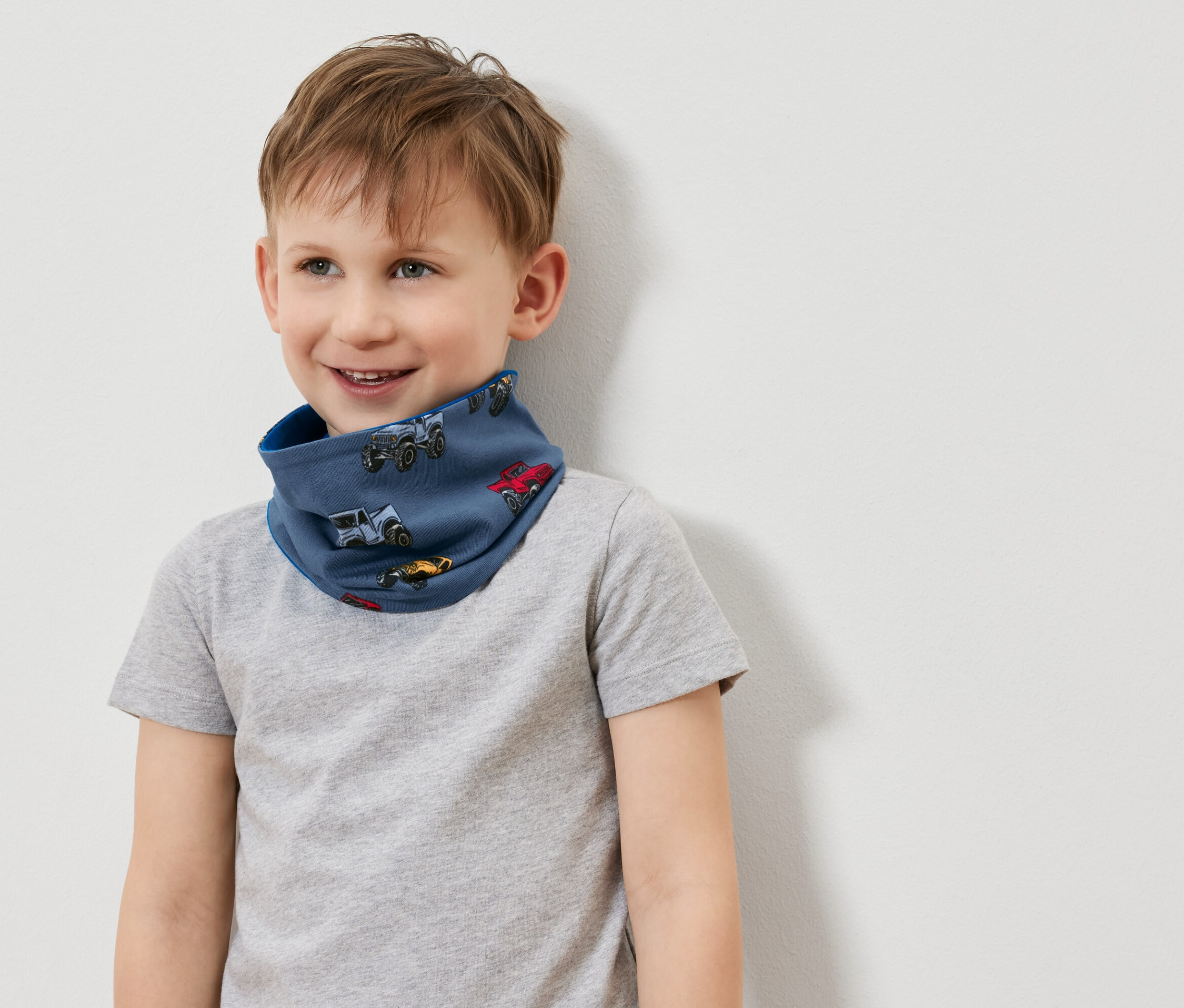 Chlapec s úsměvem nosí modrý nákrčník s potiskem autíček a šedé tričko.