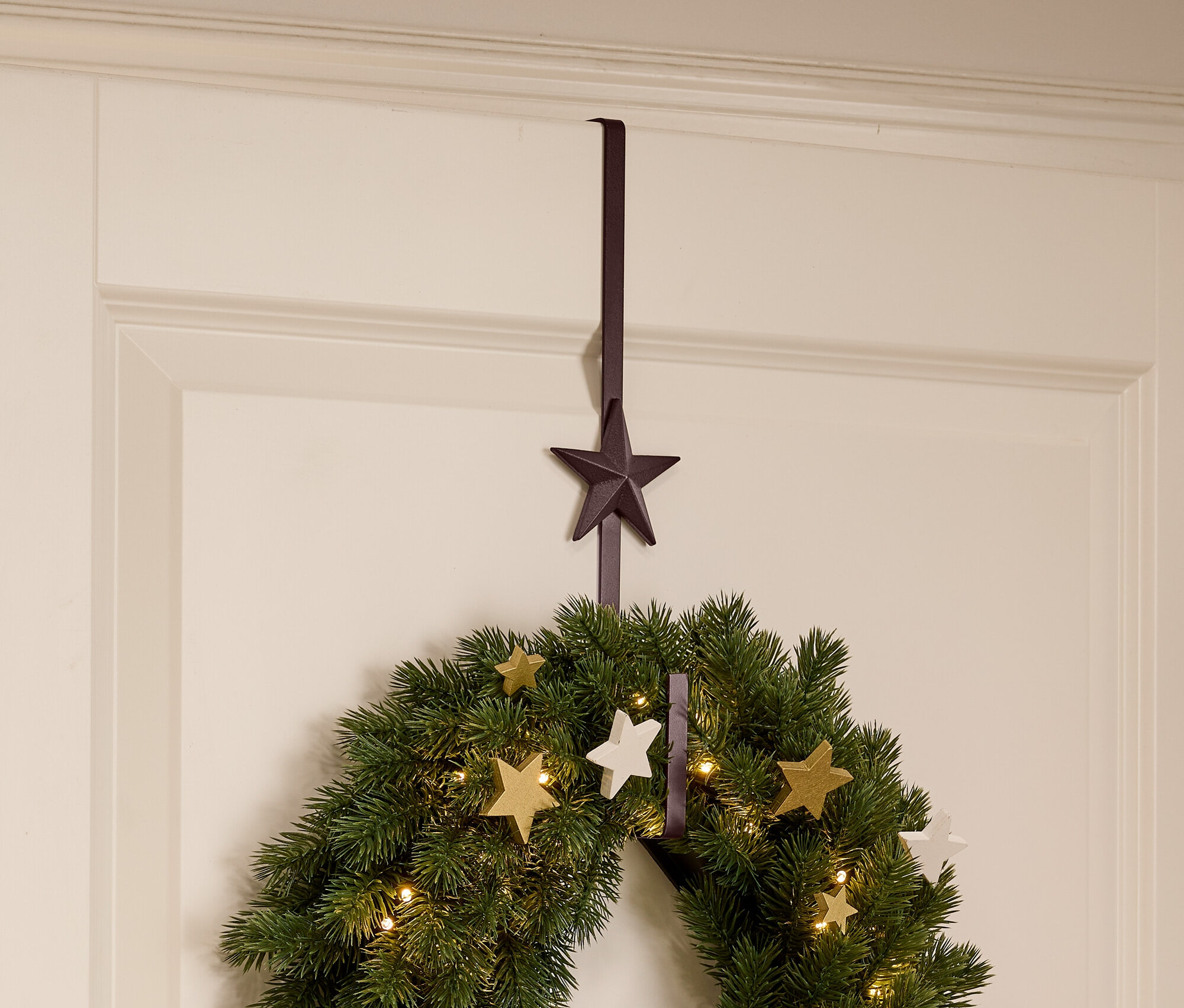 Zelený vánoční věnec s hvězdami a světly visí na bílých dveřích pomocí hnědého věšáku ve tvaru hvězdy.