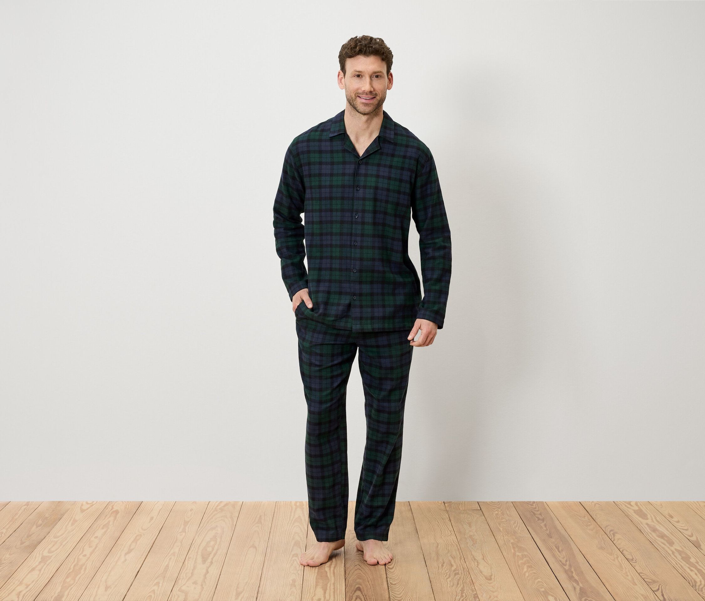 Muž v kostkovaném pyžamu stojí na dřevěné podlaze před bílou stěnou.