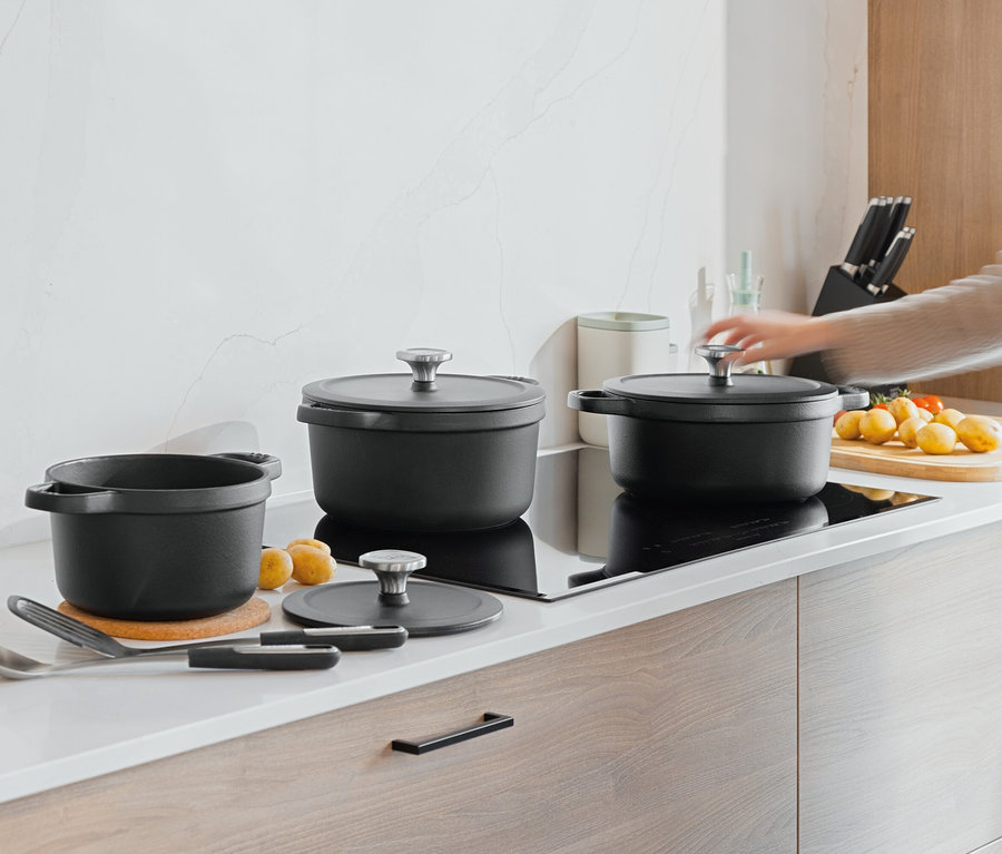 Na kuchyňské lince stojí tři černé hrnce, vedle nich leží brambory a rajčata. Ruka se natahuje k poklici jednoho z hrnců.