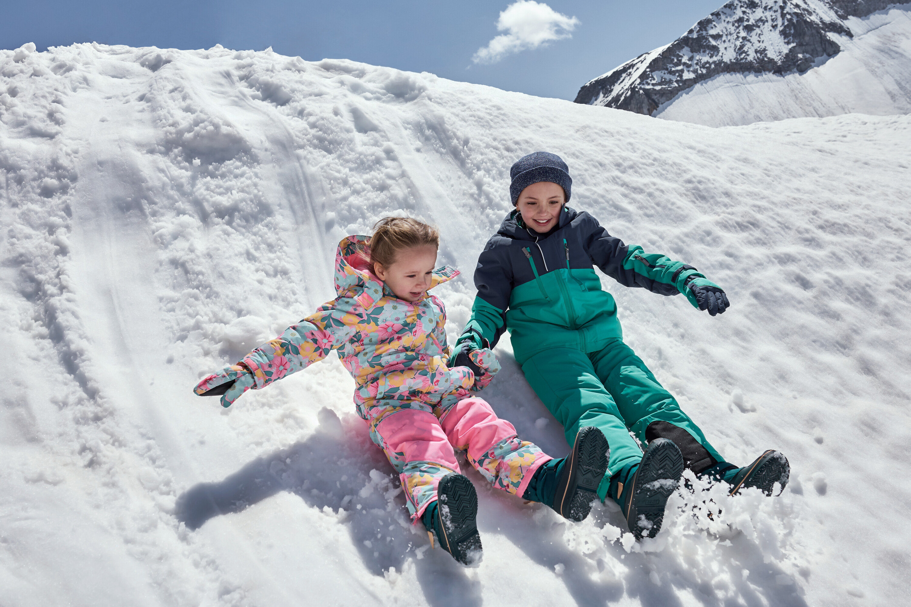 Děti sjíždějí zasněžený kopec v barevných zimních kombinézách.