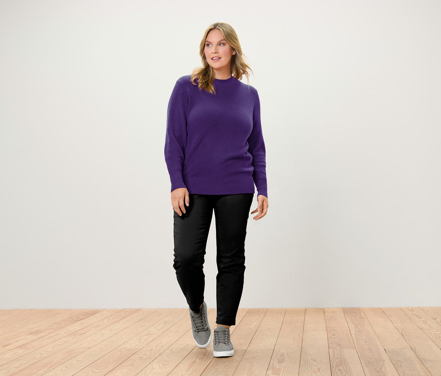 Žena kráčí po dřevěné podlaze a má na sobě fialový svetr, černé kalhoty a šedé tenisky.