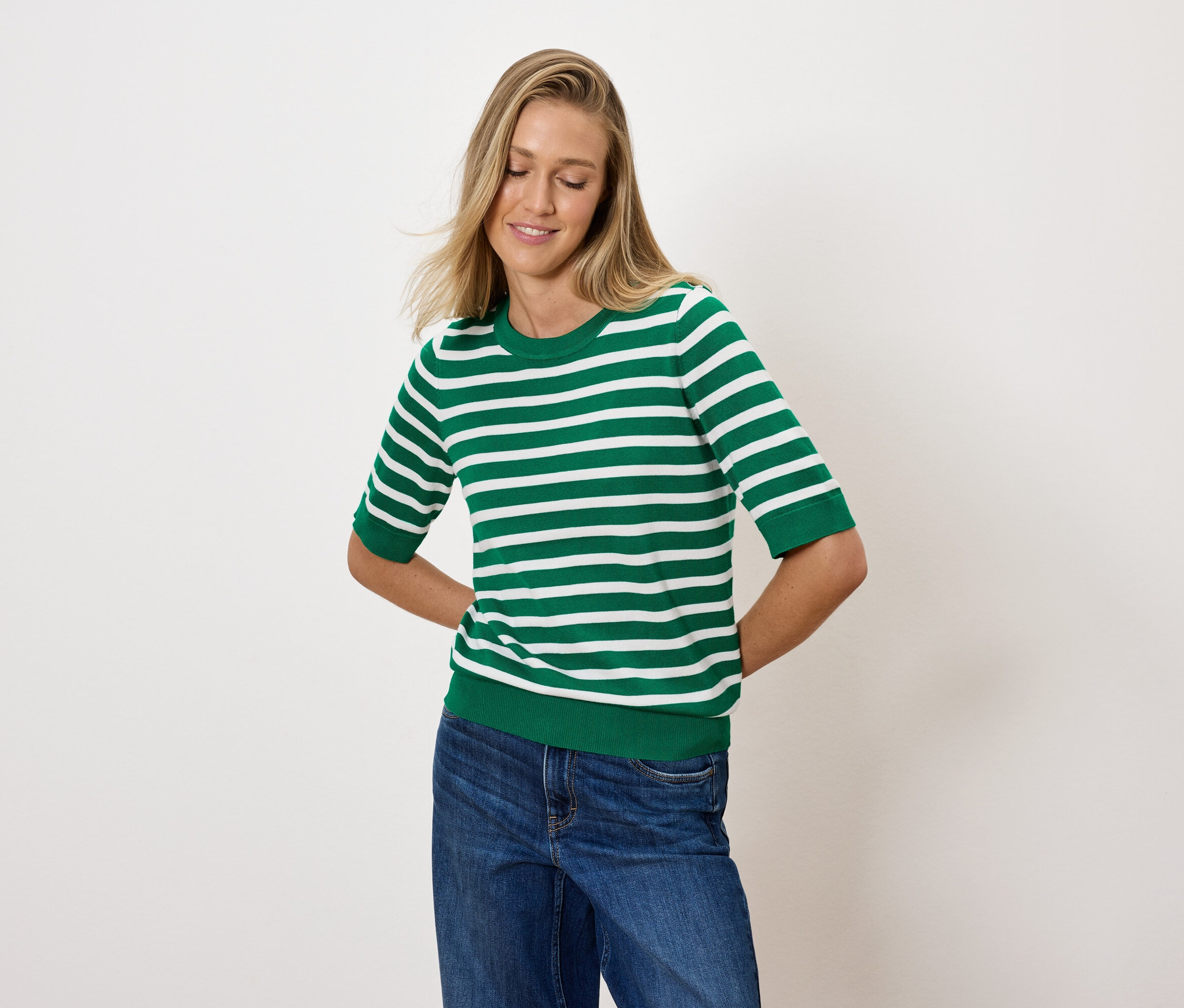 Žena s blond vlasy pózuje v zelenobílém pruhovaném topu a modrých džínách.