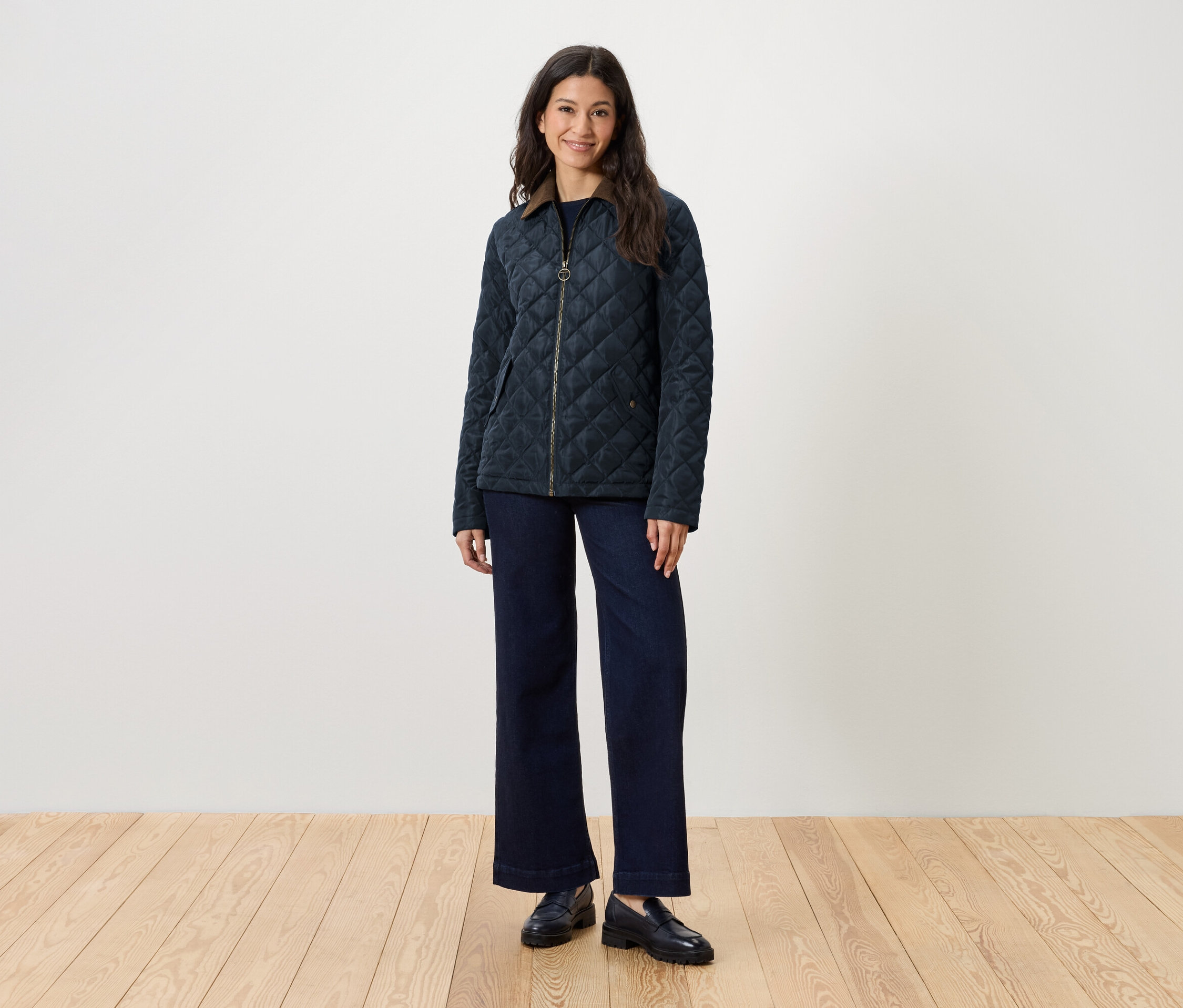 Žena stojí a má na sobě prošívanou bundu, džíny a mokasíny.