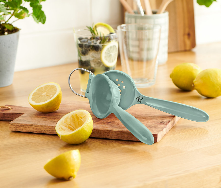 Modrý lis na citrony leží na dřevěném prkénku vedle rozpůlených citronů. V pozadí je sklenice s citronem a borůvkami.