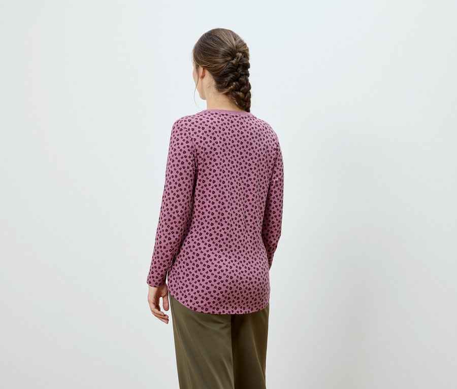 Zezadu pohled na ženu s hnědým copem, oblečenou v růžovém topu s dlouhými rukávy a tmavým potiskem, a khaki kalhotách.