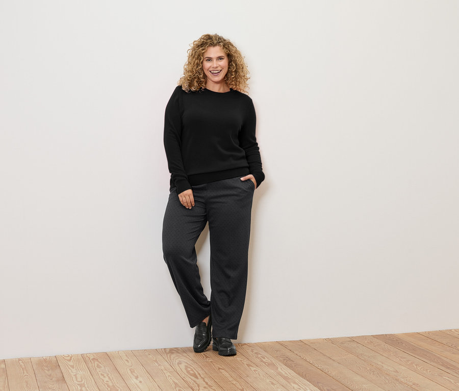 Žena s kudrnatými vlasy se opírá o zeď a má na sobě černý svetr a vzorované kalhoty.