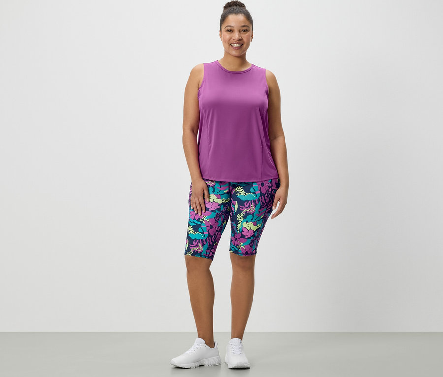 Žena stojí a má na sobě sportovní cyklistické šortky s celoplošným potiskem a tyrkysový sportovní top z 2 ks setu.