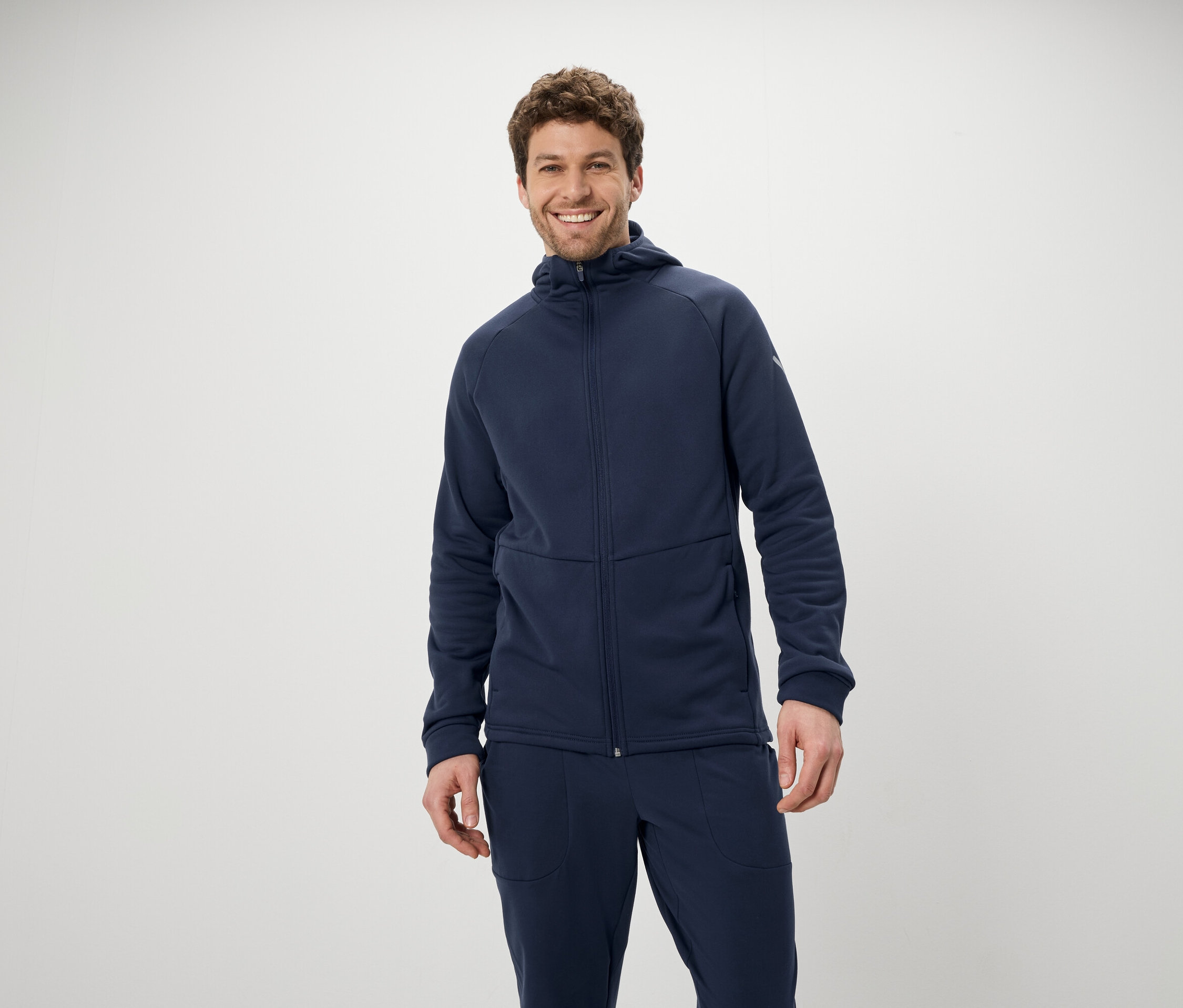 Usmívající se muž pózuje v modré soupravě s kapucí a kalhotami na bílém pozadí.