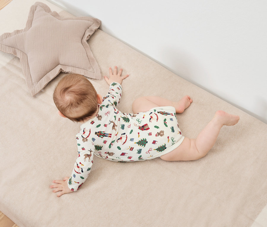 Dítě leží na břiše na dece s vánočním potiskem vedle béžového hvězdicového polštáře.