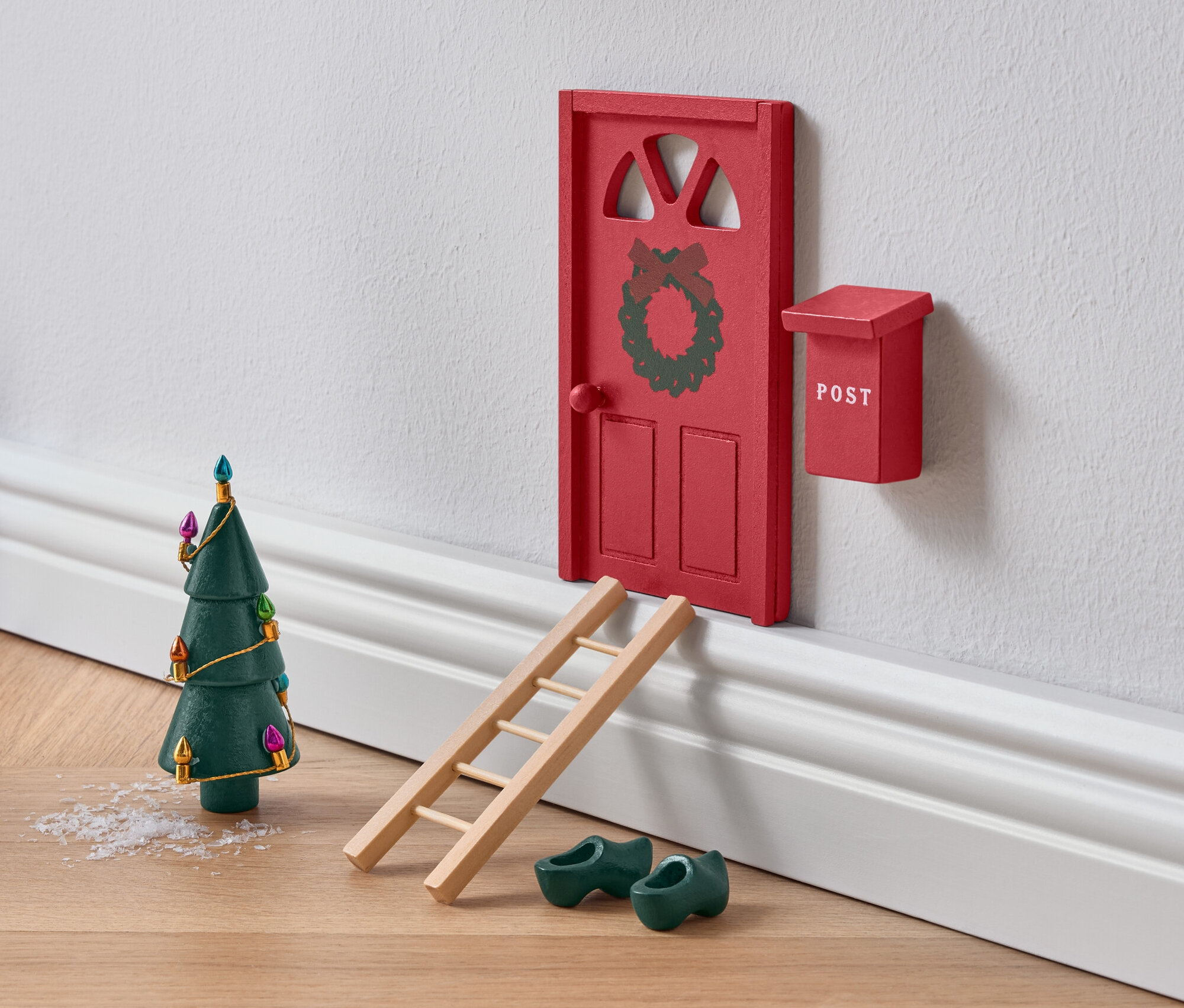 Miniaturní červené dveře zdobené věncem s žebříkem, zelenými dřeváky a vánočním stromkem.