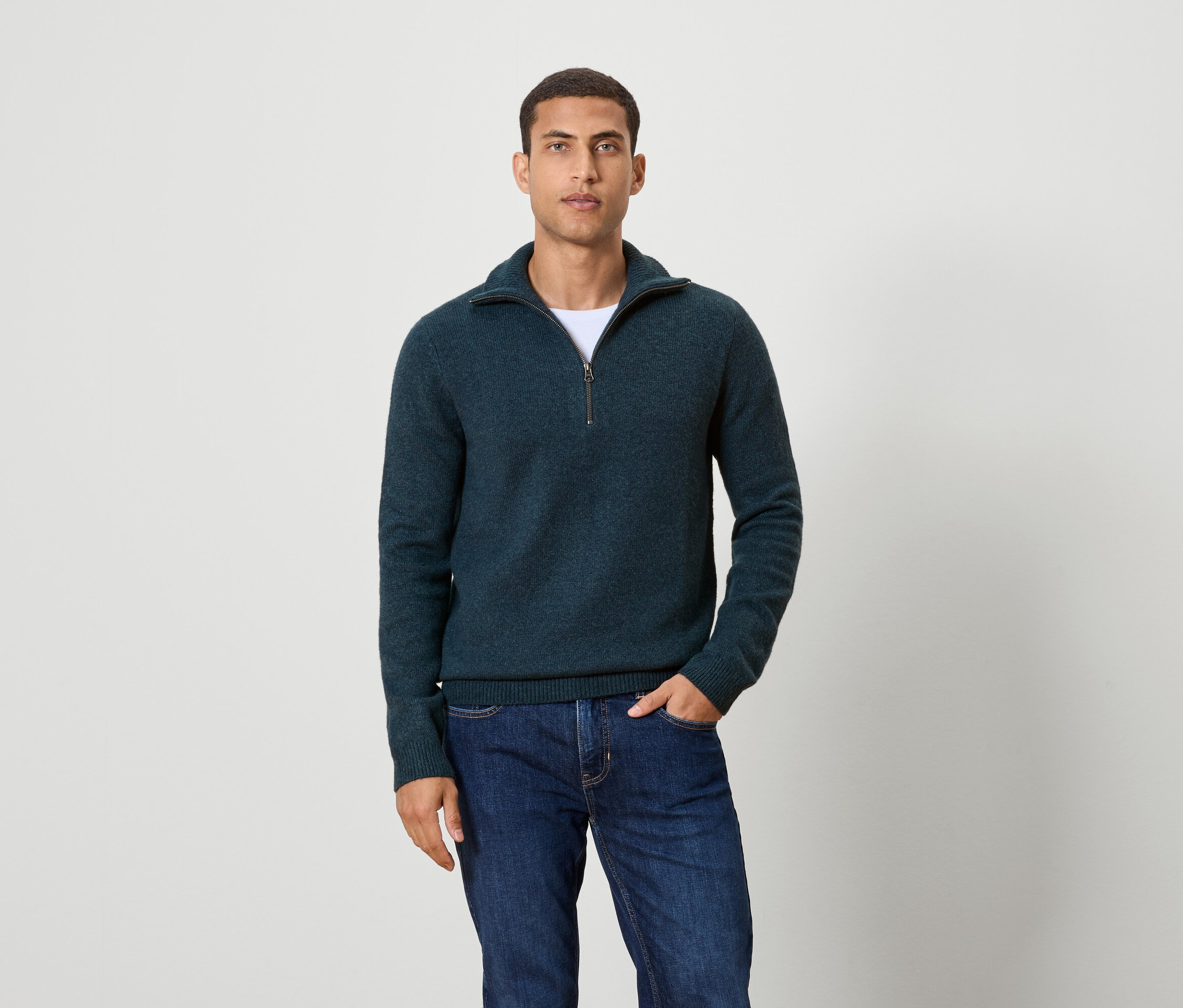 Muž pózuje na pozadí a má na sobě tmavě modrý svetr na zip a džíny.