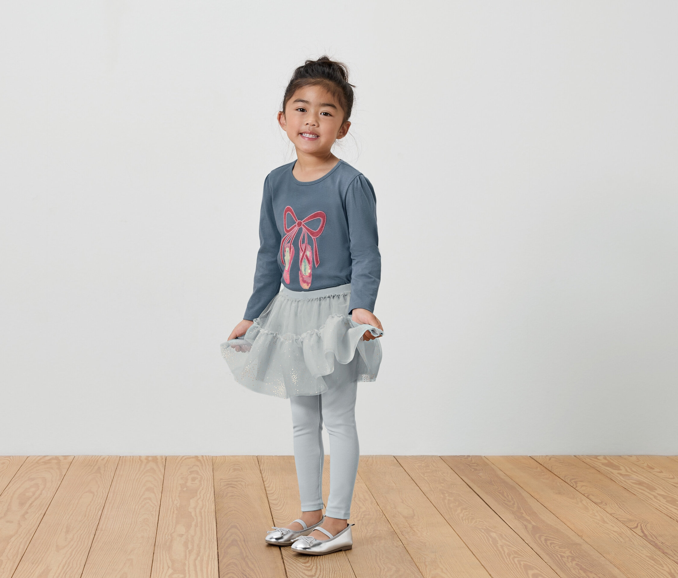 Dívka v šedém tričku s baletními střevíci, sukni a legínách.