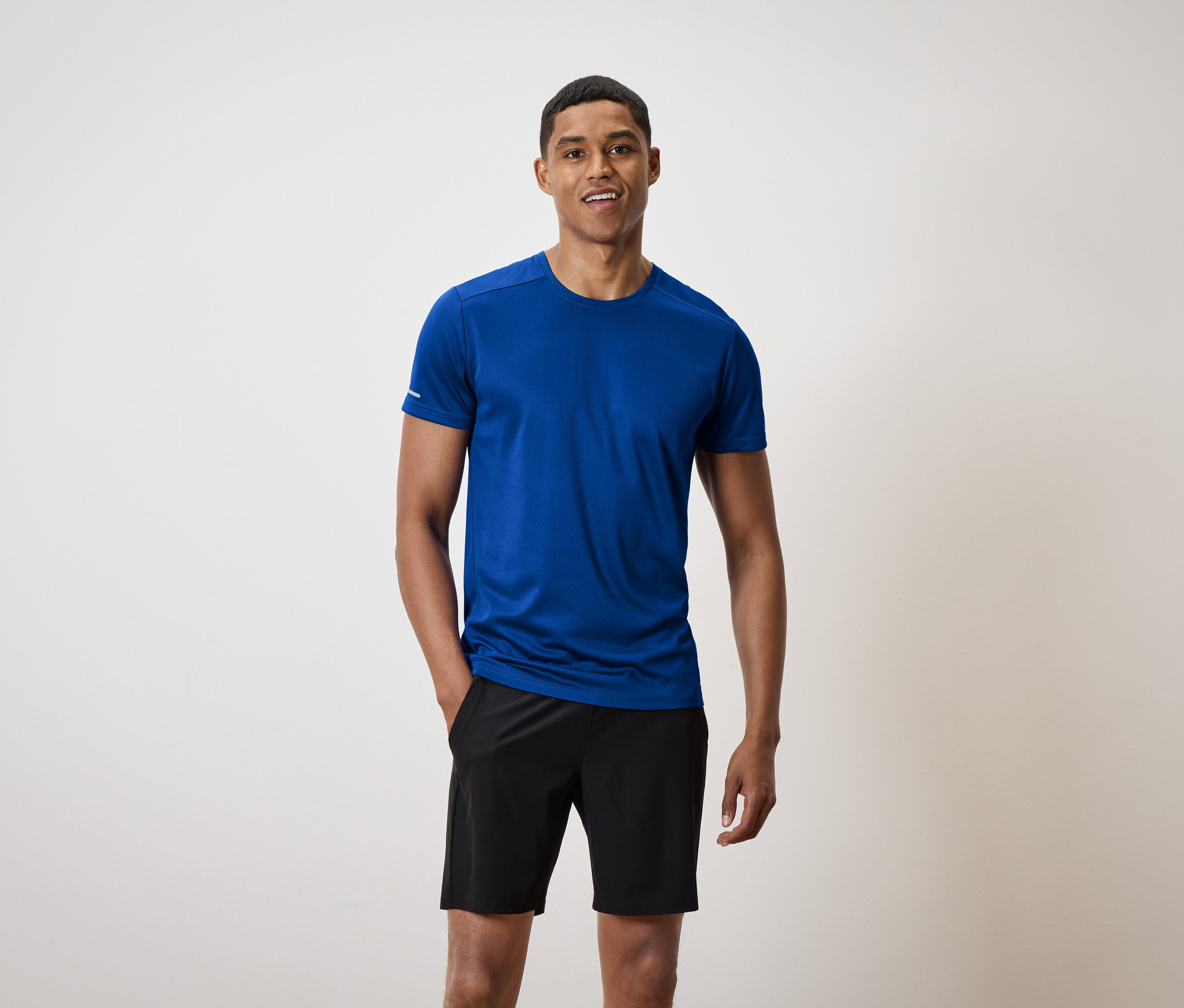 Muž s krátkými vlasy stojí a usmívá se, má na sobě modré tričko a černé šortky.