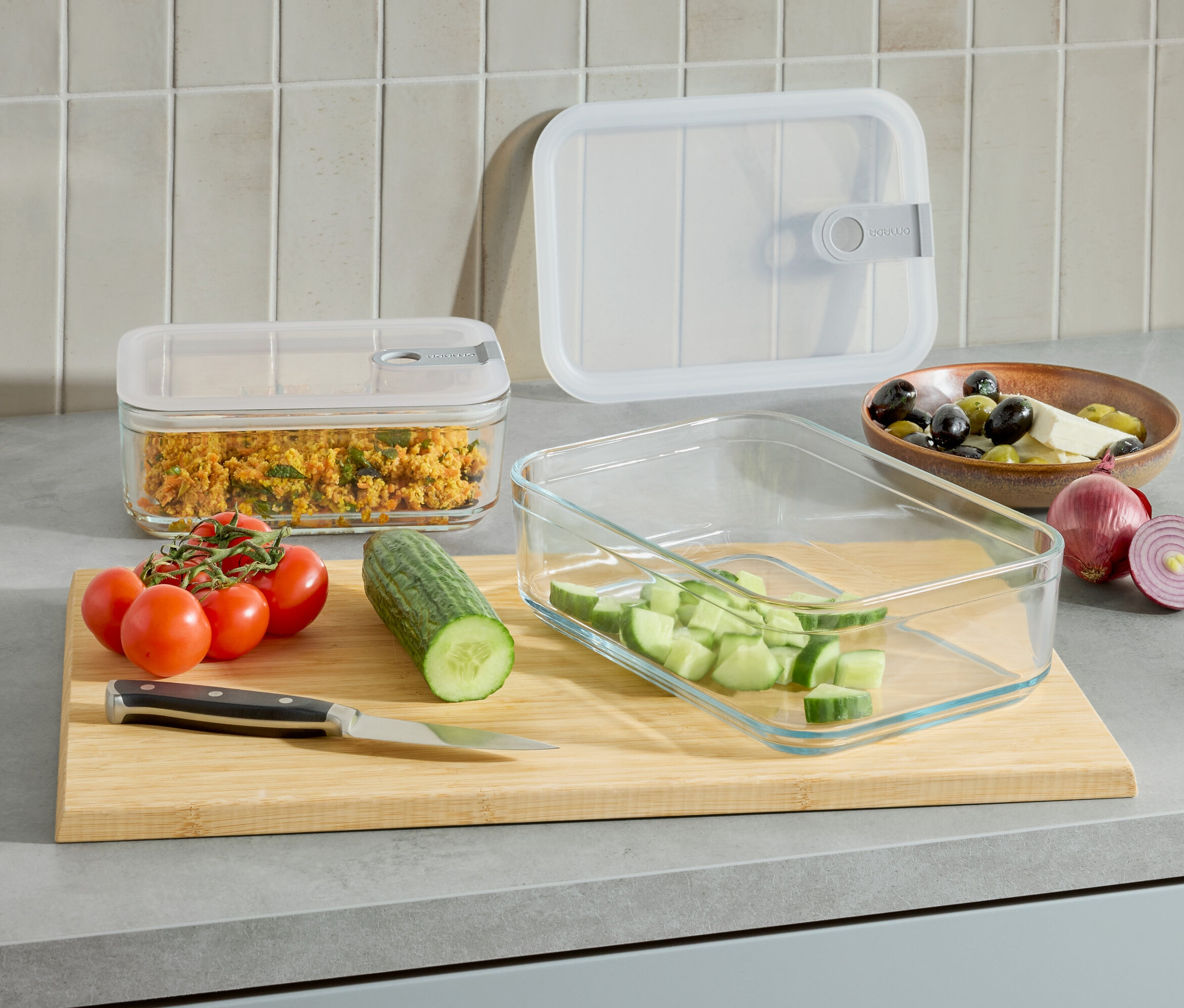 Na lince jsou 2 skleněné nádoby na potraviny, jedna s jídlem, druhá s nakrájenou okurkou. Dále jsou tam rajčata, okurka, nůž a mísa s olivami a sýrem.