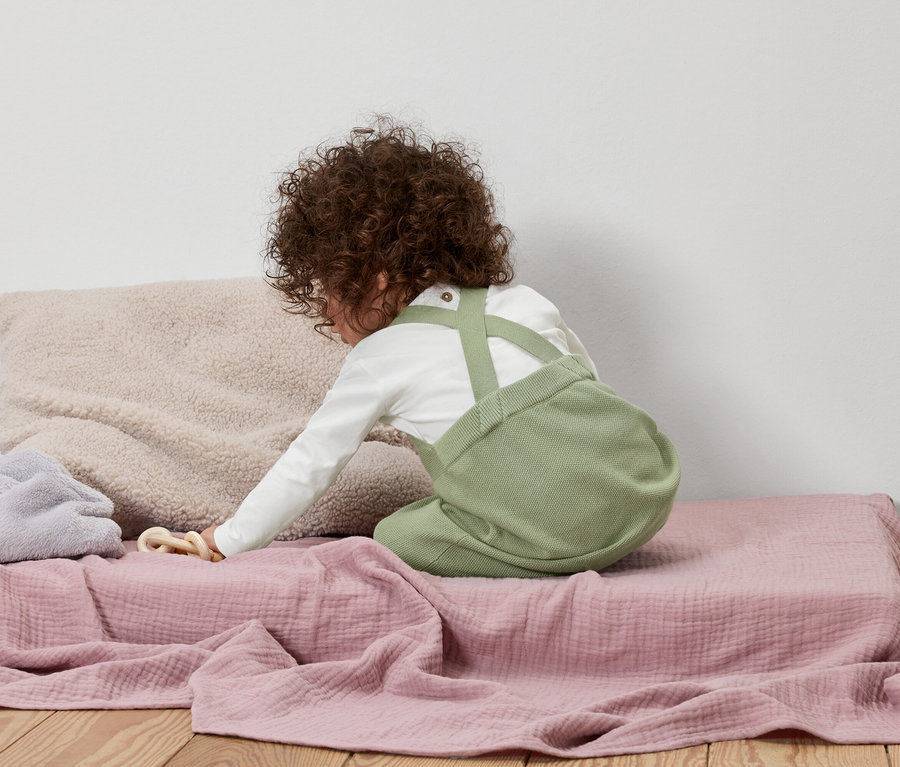 Dítě s kudrnatými vlasy klečí na růžové dece, oblečené v zelených lacláčích a tričku s dlouhým rukávem.