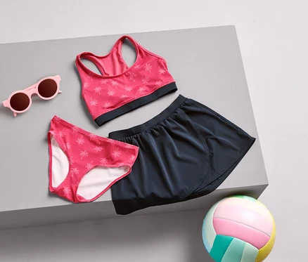 Růžové sluneční brýle, bikiny kalhotky s palmovým vzorem, sportovní podprsenka a tmavě modrá sukně leží na šedém povrchu, spolu s barevným volejbalovým míčem.