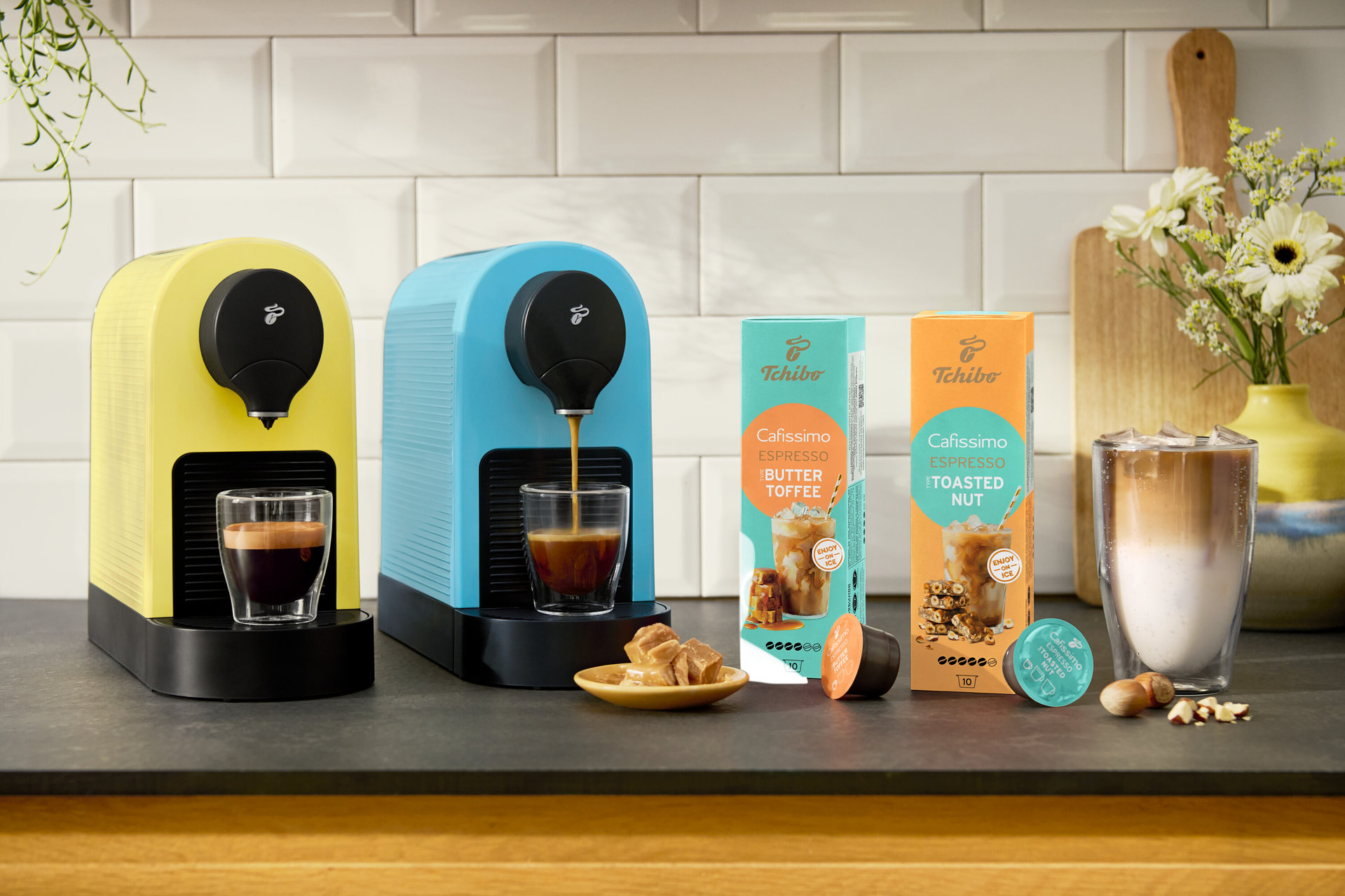 Dva kávovary Cafissimo pure+ ve žluté a modré barvě připravují espresso. Viditelné jsou také kapsle Cafissimo Flavoured Espresso – Toasted Nut.