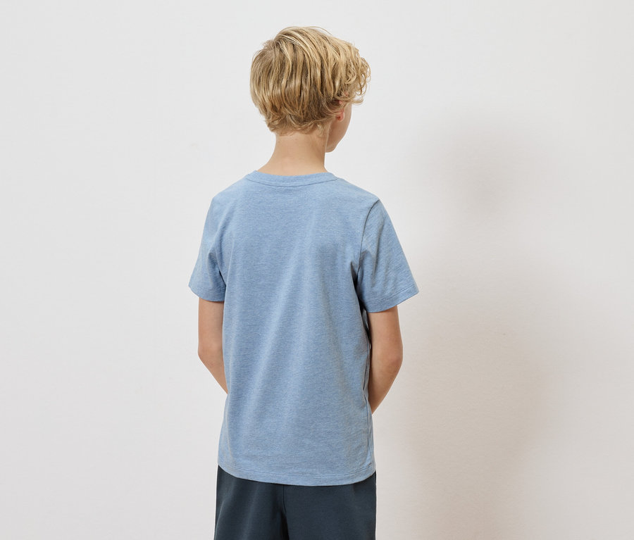 Pohled zezadu na dítě s blond kudrnatými vlasy, oblečené ve světle modrém tričku a tmavě modrých šortkách na bílém pozadí.