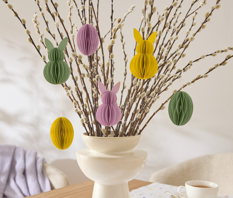 Dekorativní velikonoční větvičky s visícími papírovými ozdobami ve tvaru zajíčků a vajíček v žluté, zelené a fialové barvě ve váze.