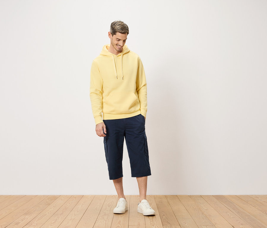 Muž ve žluté mikině s kapucí a modrých cargo šortkách stojící na dřevěné podlaze.