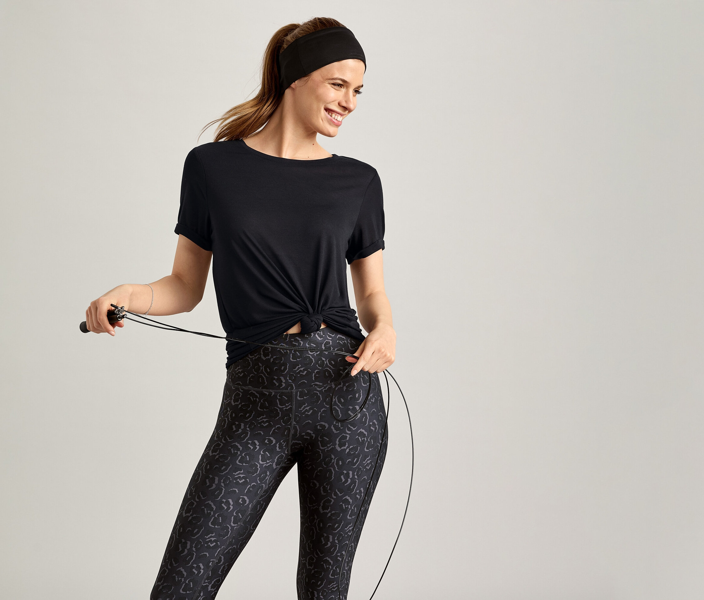 Žena s čelenkou a v černém sportovním tričku drží švihadlo na šedém pozadí.