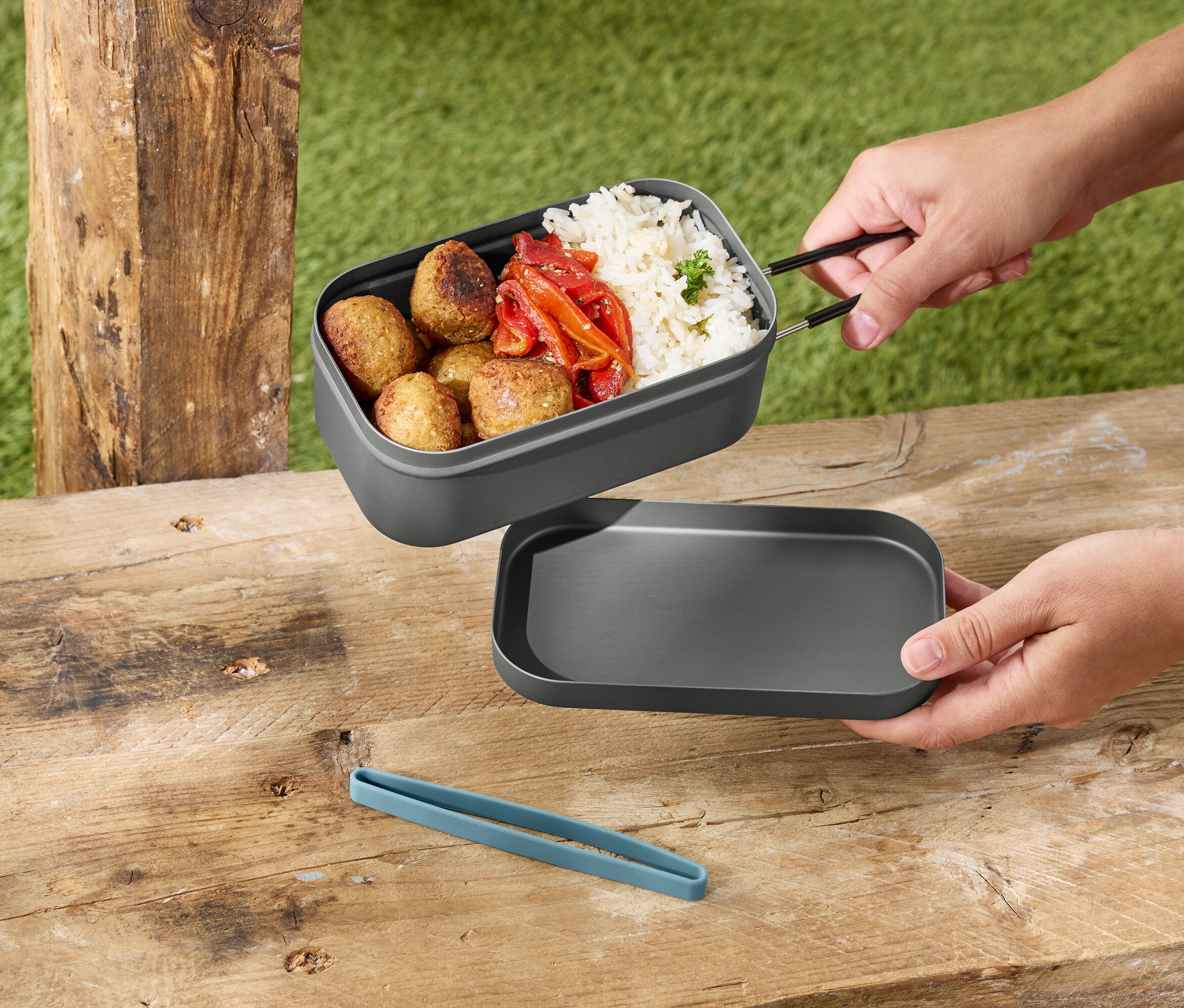 Ruce drží šedý obědový box s rýží, masovými kuličkami a paprikou nad šedým víkem na dřevěném stole.