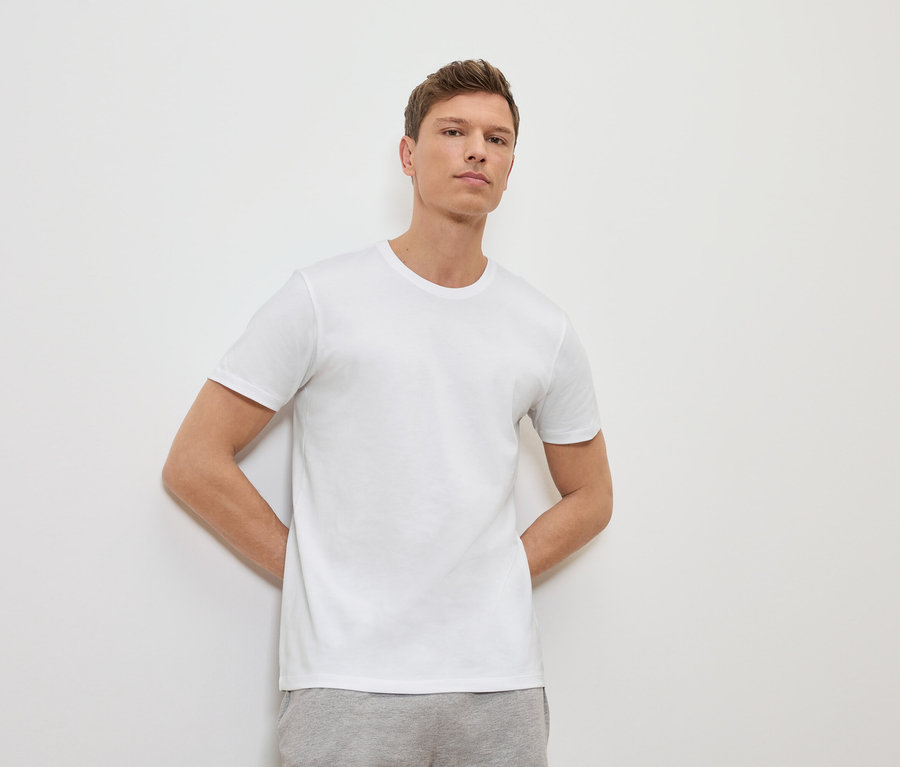 Muž se opírá o bílou stěnu a má na sobě bílé tričko a šedé tepláky.