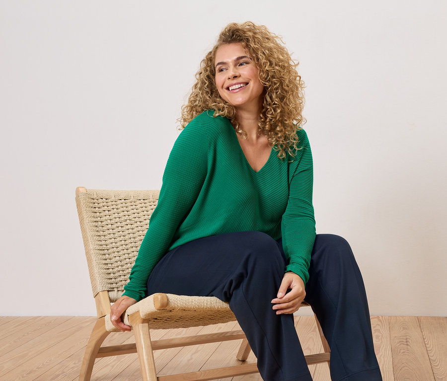 Žena s kudrnatými vlasy sedí na židli a má na sobě zelený svetr a modré kalhoty.