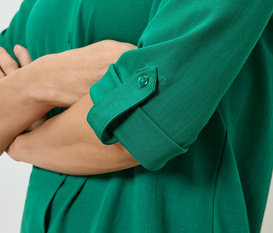Detail osoby se zkříženýma rukama, která má na sobě zelenou košili s knoflíky a ohrnutými rukávy.