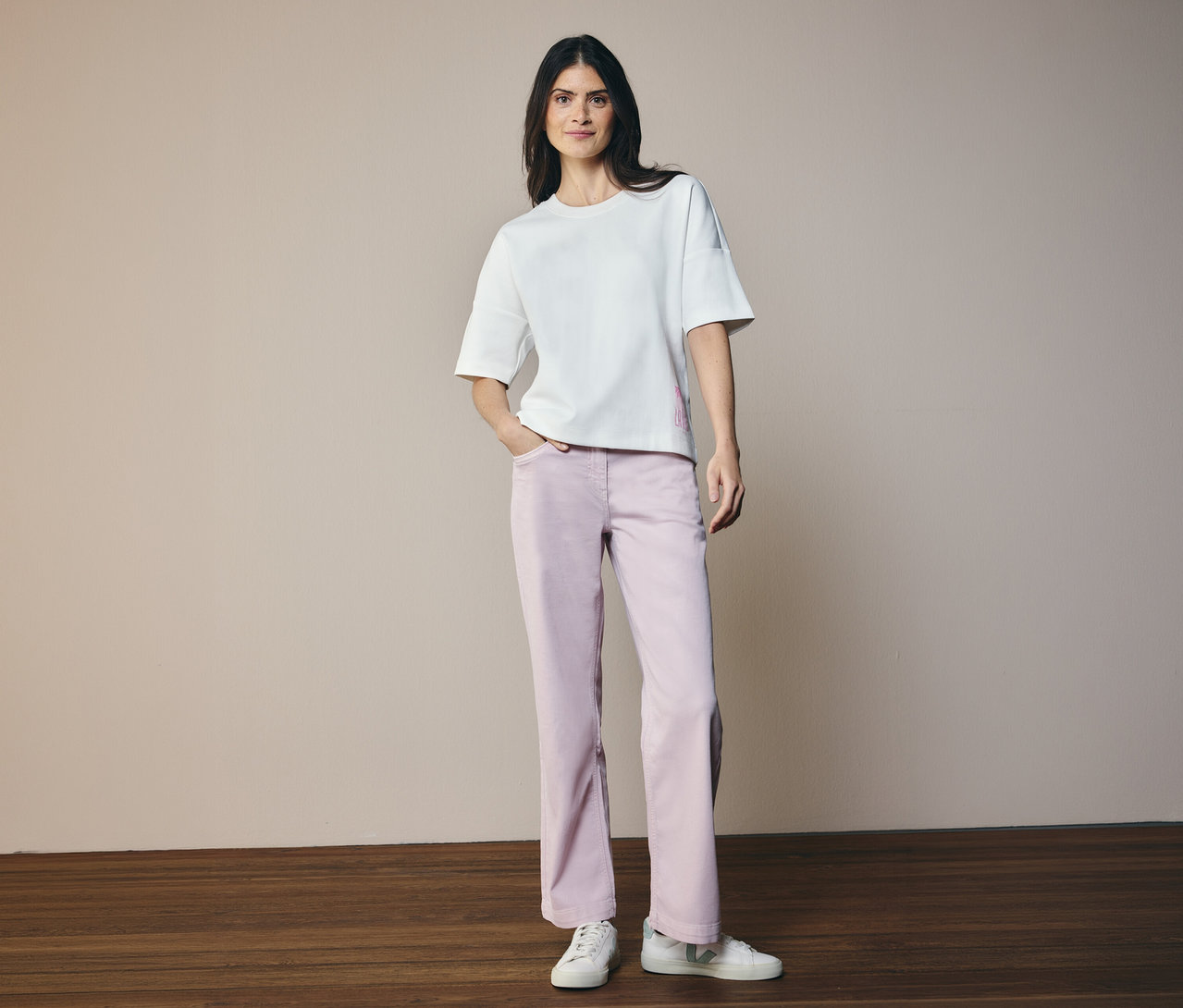 Žena v bílém tričku a džíny CECIL »Neele«. Stojí na dřevěné podlaze před béžovým pozadím.