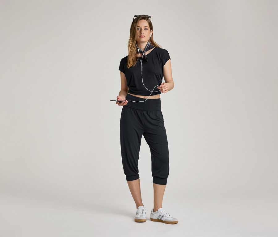 Žena se sluchátky a mobilem. Má na sobě 3/4 kalhoty na jógu, černý top a tenisky.