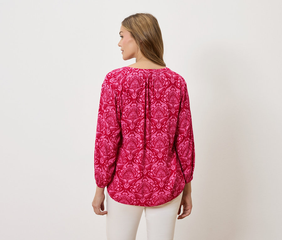 Pohled zezadu na ženu s blond vlasy, která má na sobě růžovou halenku s dlouhým rukávem se vzorem a bílé kalhoty.