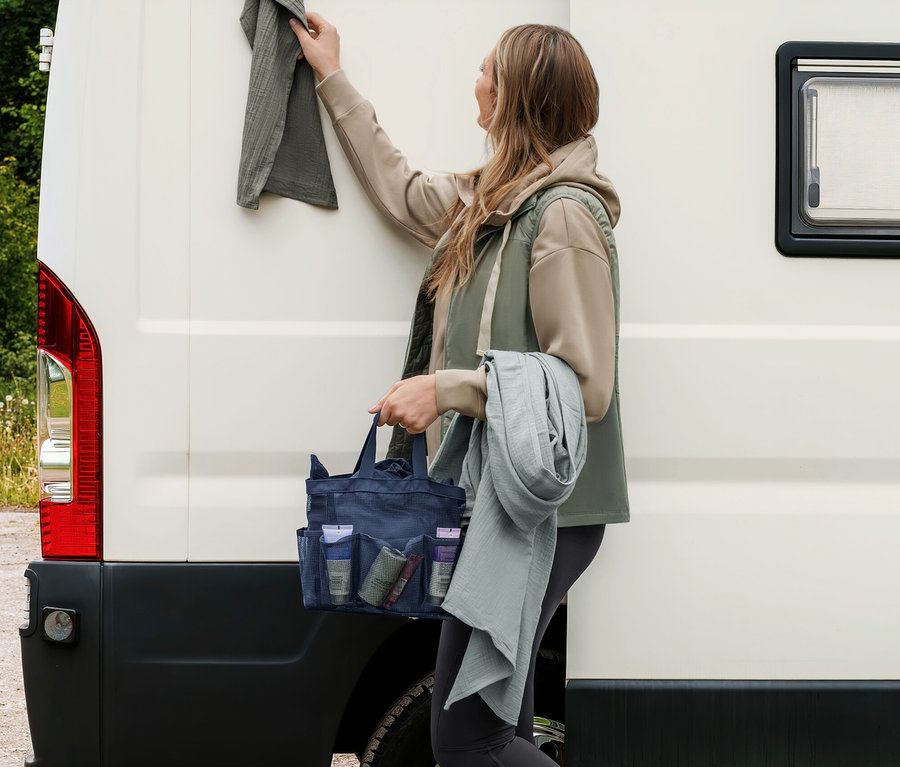 Žena věší šedozelený mušelínový ručník na karavan, drží tmavě modrou tašku do sprchy a další světle šedozelený mušelínový ručník.