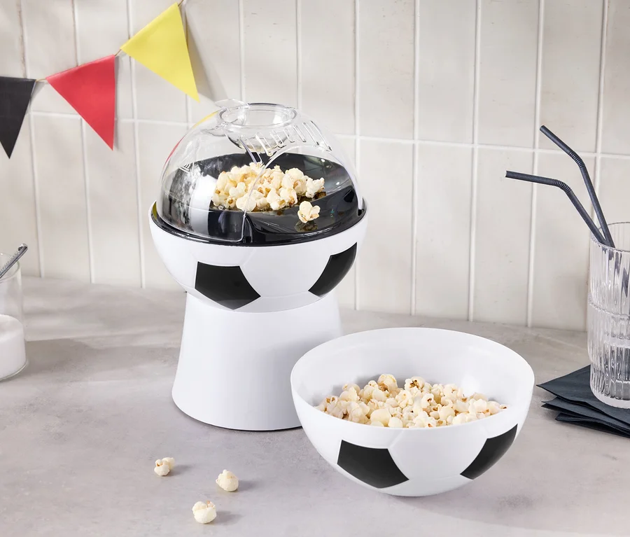 Stroj na popcorn ve tvaru fotbalového míče stojí vedle misky popcornu a sklenice s brčky.