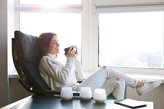 Relax v křesle: Žena oblečená v bílém sedí před oknem v relaxačním křesle z černé kůže a v ruce drží šálek čaje.