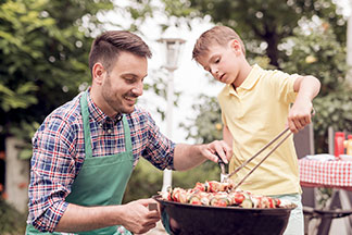 Mladý muž se zelenou zástěrou klečí vedle černého, kulatého grilu, zatímco jeho syn kleštěmi otáčí masové špízy.