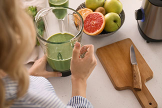 Kuchyňské spotřebiče v akci: V kuchyni stojí žena a v ruce drží mixér se zeleným smoothie – vedle je miska s ovocem a dřevěné prkénko na krájení s nožem.