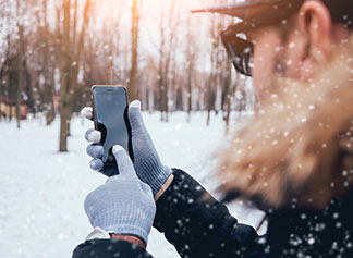 Rukavice pro muže s důležitými detaily: Muž stojí v zasněžené zimní krajině a s rukavicemi na rukou obsluhuje svůj smartphone.