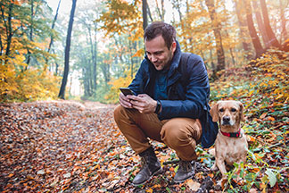 Muž v modré přechodné bundě a nepromokavém pánském vysokém botníku klečí v podřepu v podzimním lese a obsluhuje svůj chytrý telefon, vedle něj leží jeho pes.