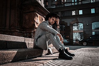 Pánské vysoké boty pro business look: mladý muž v černých botách, kostkovaných kalhotách, kostkovaném saku a klobouku sedí venku na kamenném schodišti.