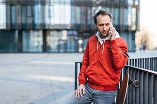 Pánská sportovní bunda pro každodenní použití: Mladý muž v červené sportovní bundě telefonuje, vedle sebe má položený longboard.