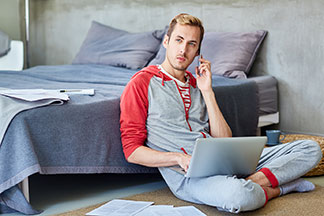 Mladý muž oblečený do domácí soupravy pracuje vsedě na zemi – opřený o postel – na laptopu a přitom telefonuje.